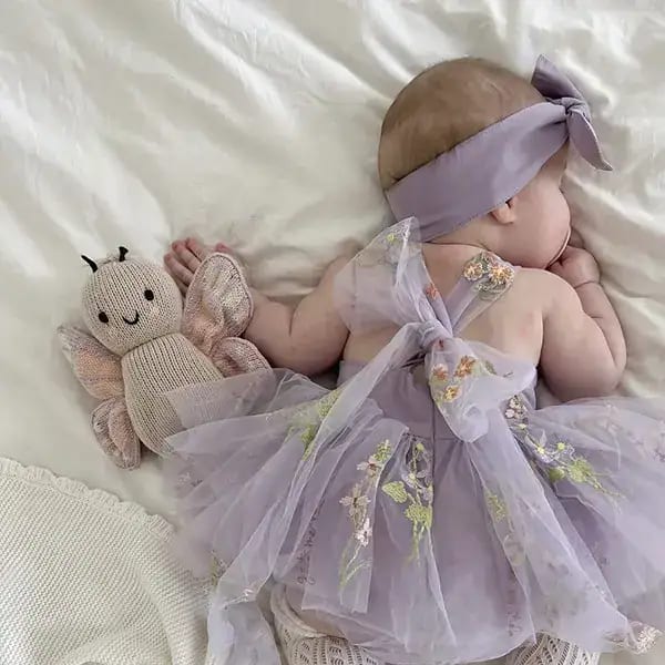 Bebé durmiendo con vestido de encaje y diadema en tonos lilas, acompañada de un muñeco mariposa.